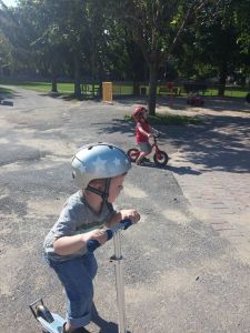 At the park, Liam plays on his scooter with his friend riding his bike in the background